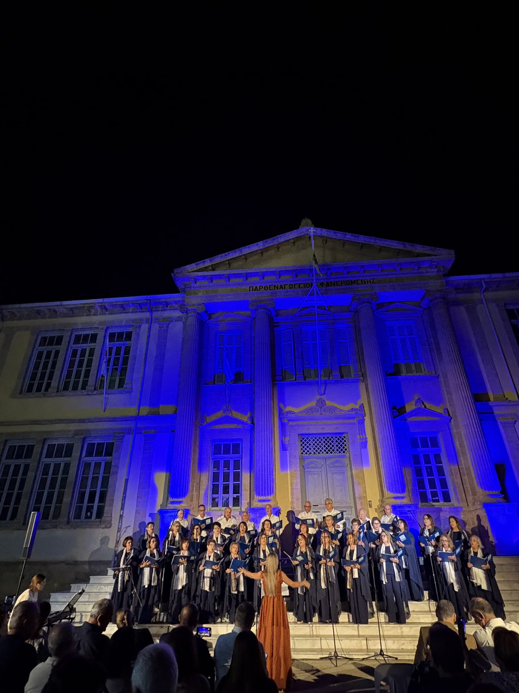Choir performing an open-air evening concert outside a neoclassical building lit blue