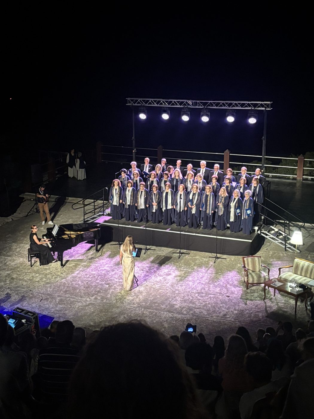 Choir performing an open-air night concert with piano accompaniment under stage lights